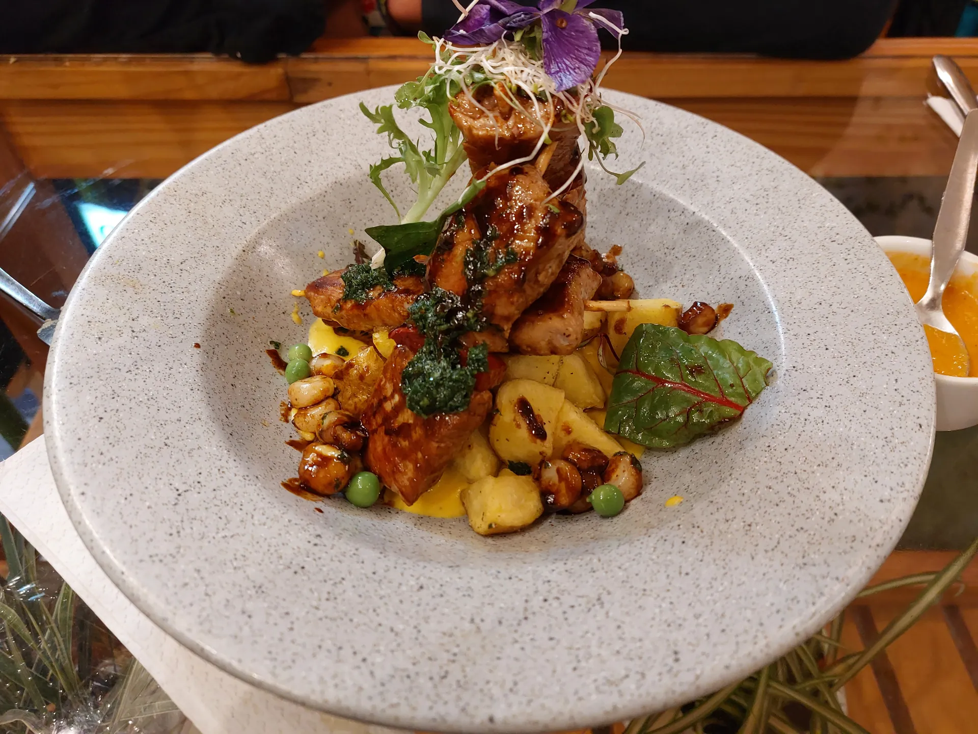 Grilled alpaca skewers on potatoes and choclo with herb sauce, aji cream, and greens in a shallow bowl.
