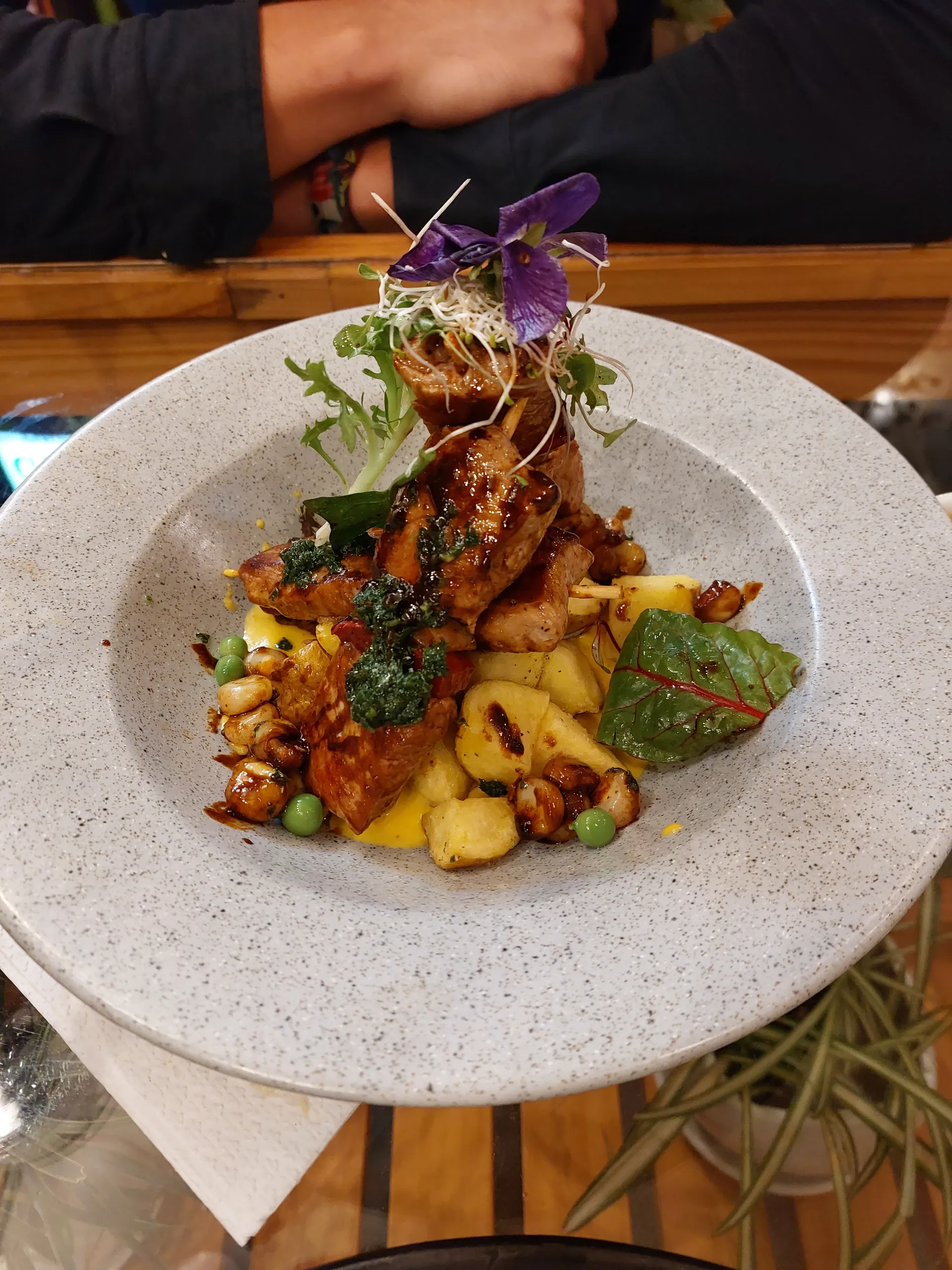 Alpaca anticuchos on skewers over potatoes and corn with glossy glaze and microgreens.