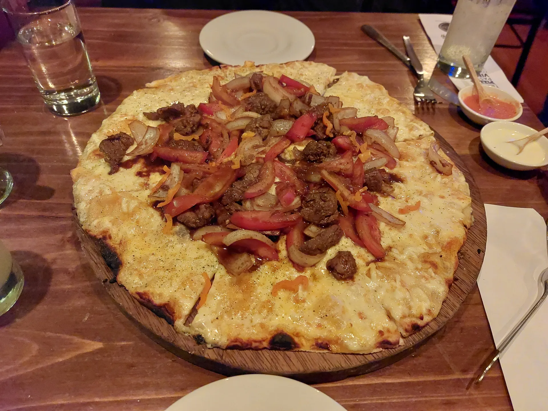 Thin-crust pizza topped with lomo saltado-style stir-fry, cheese, and dipping sauces on a wooden board.