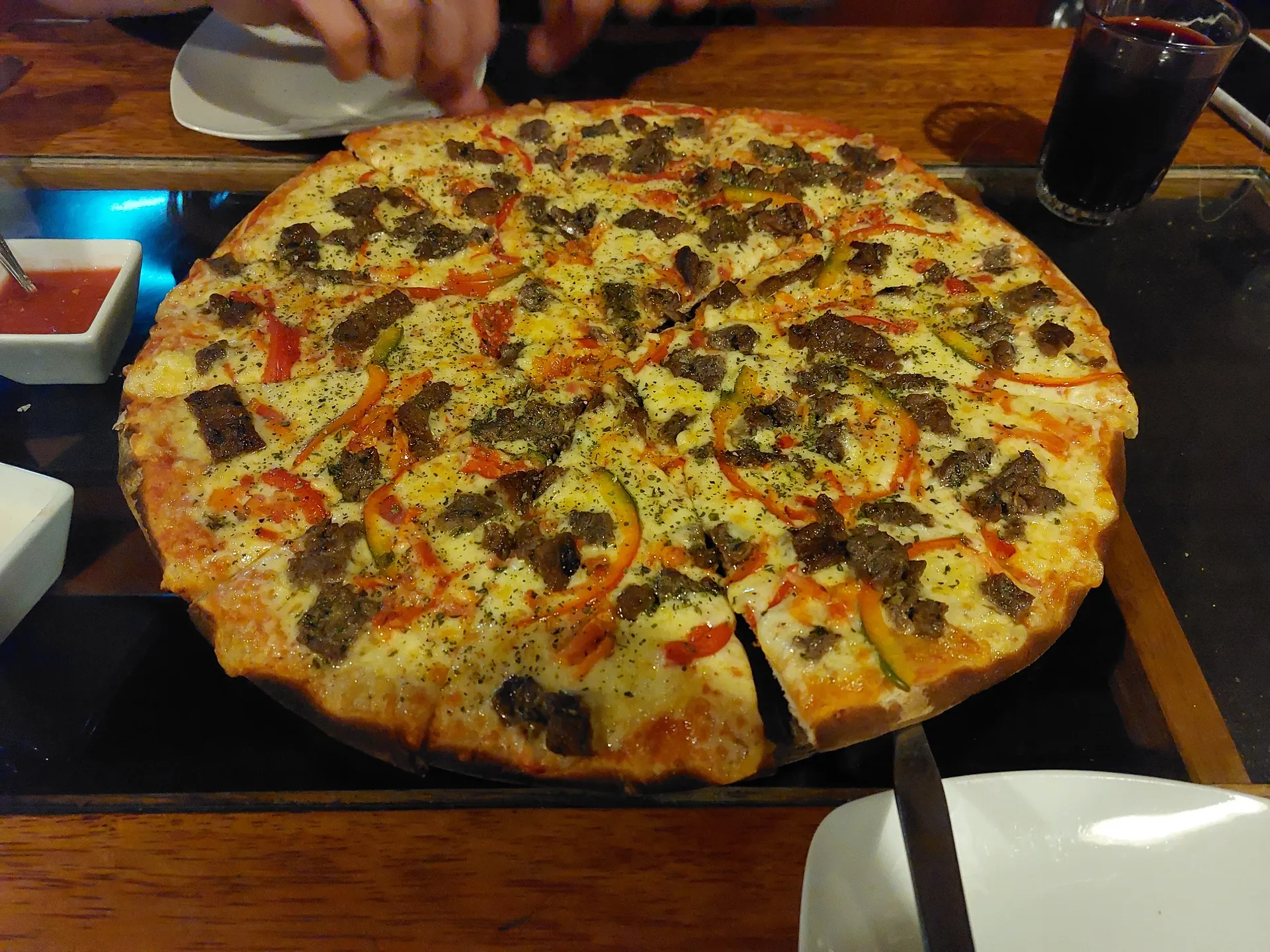 Alpaca-topped pizza with bell peppers and herbs beside a glass of chicha morada and ají sauces.