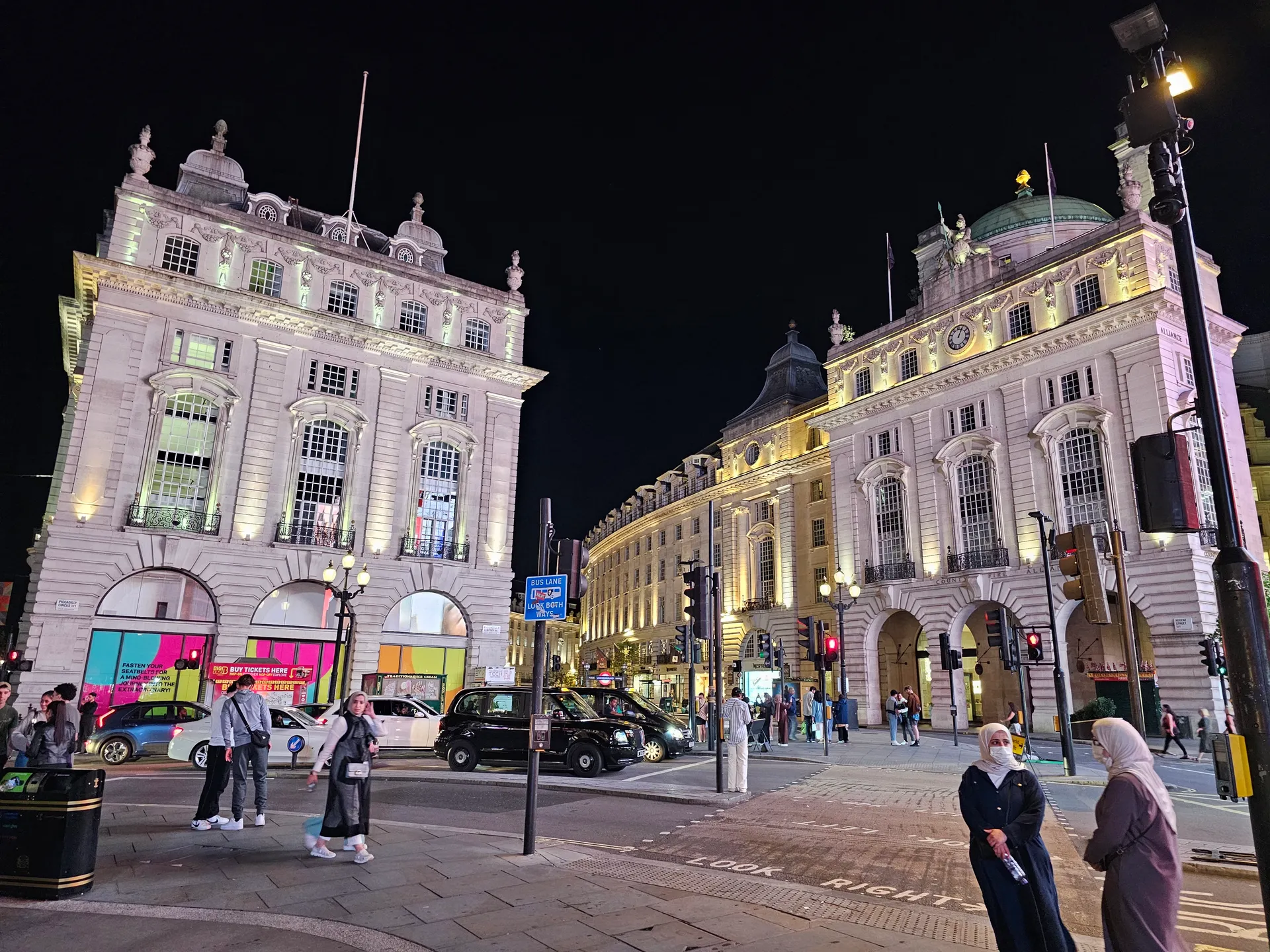 Piccadilly area at night: lit stone buildings, black taxi in the intersection, traffic lights and pedestrians.